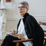 Senior woman with glasses sitting in a stylish office setting, holding a notebook and pen, signifying professionalism.